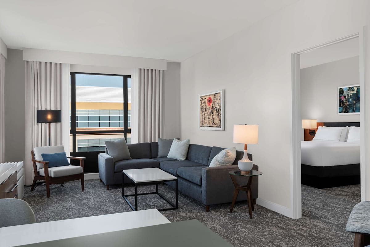 Een hotelkamer met een grijze modulaire bank, een klein tafeltje, een bed en uitzicht op de stad door grote ramen.