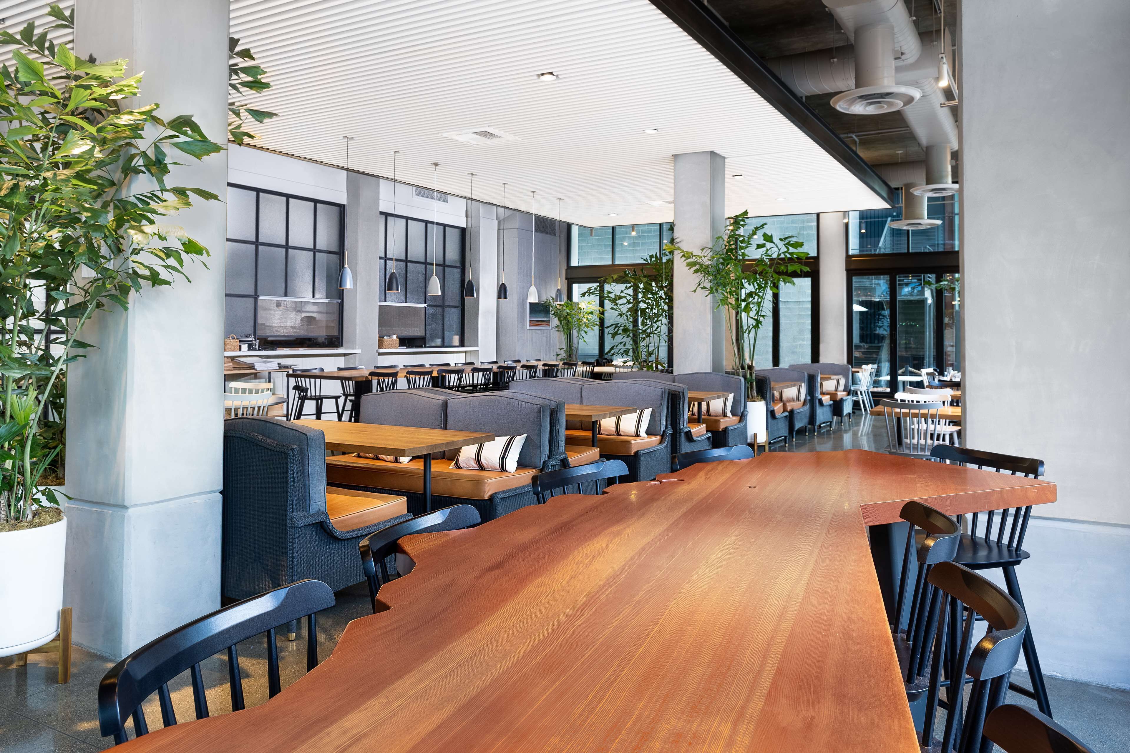 A modern indoor dining area with long wooden tables, cushioned seating, and large windows allowing natural light.