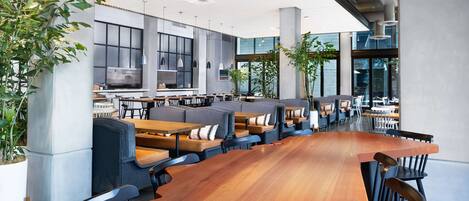 A modern indoor dining area with long wooden tables, cushioned seating, and large windows allowing natural light.