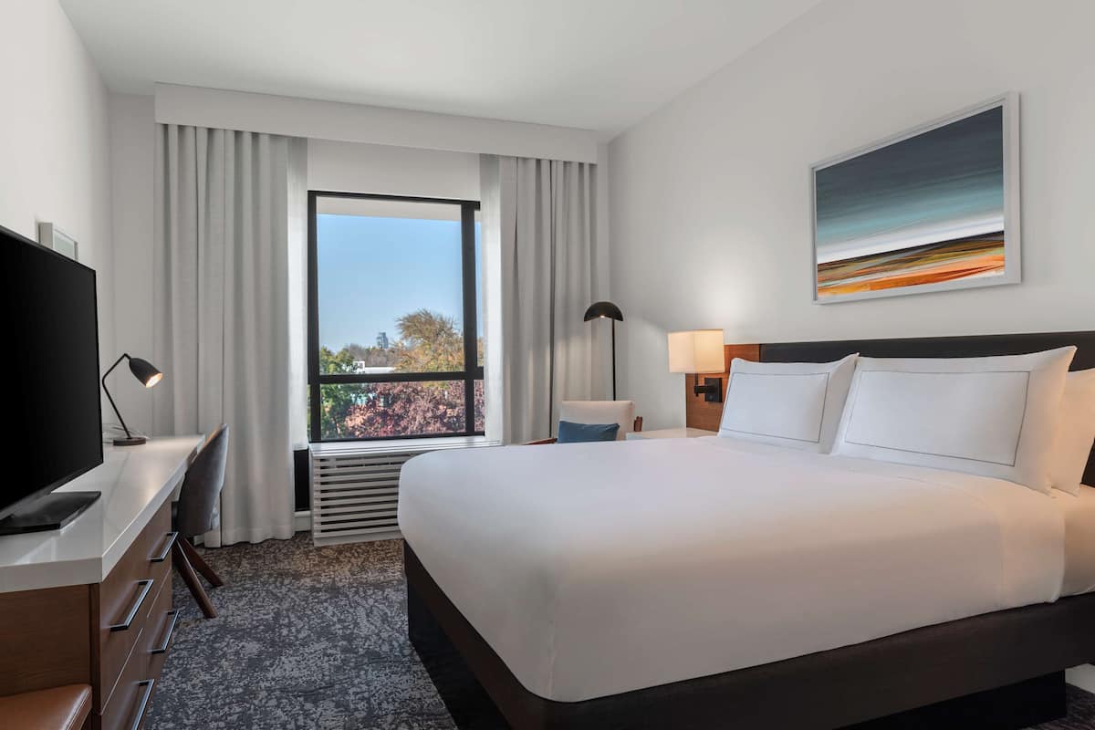 Een hotelkamer met een groot bed, een bureau met een televisie, een raam met gordijnen, een lamp en een schilderij aan de muur.