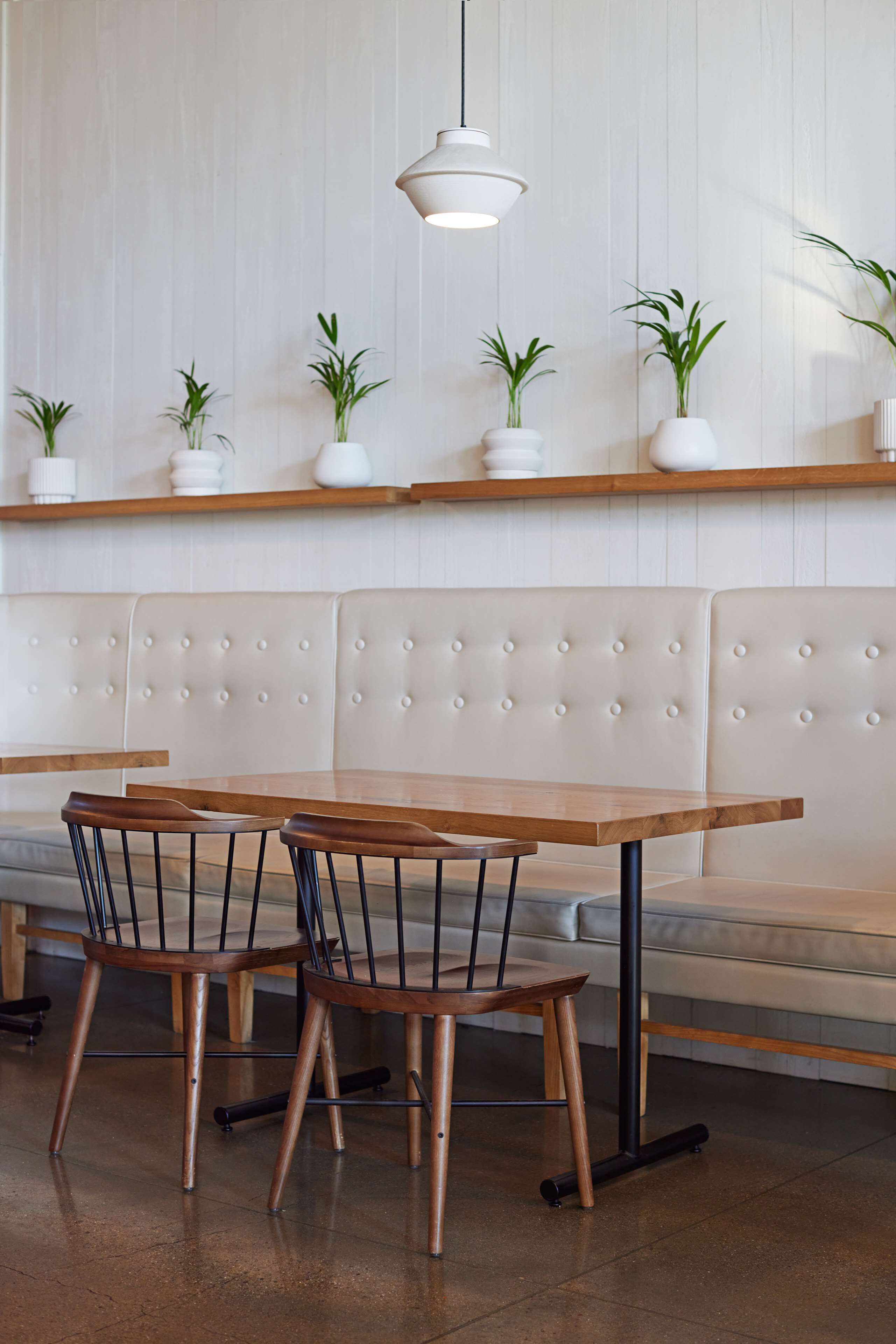 A seating area with a wooden table and chairs, a cushioned bench, and potted plants on shelves.
