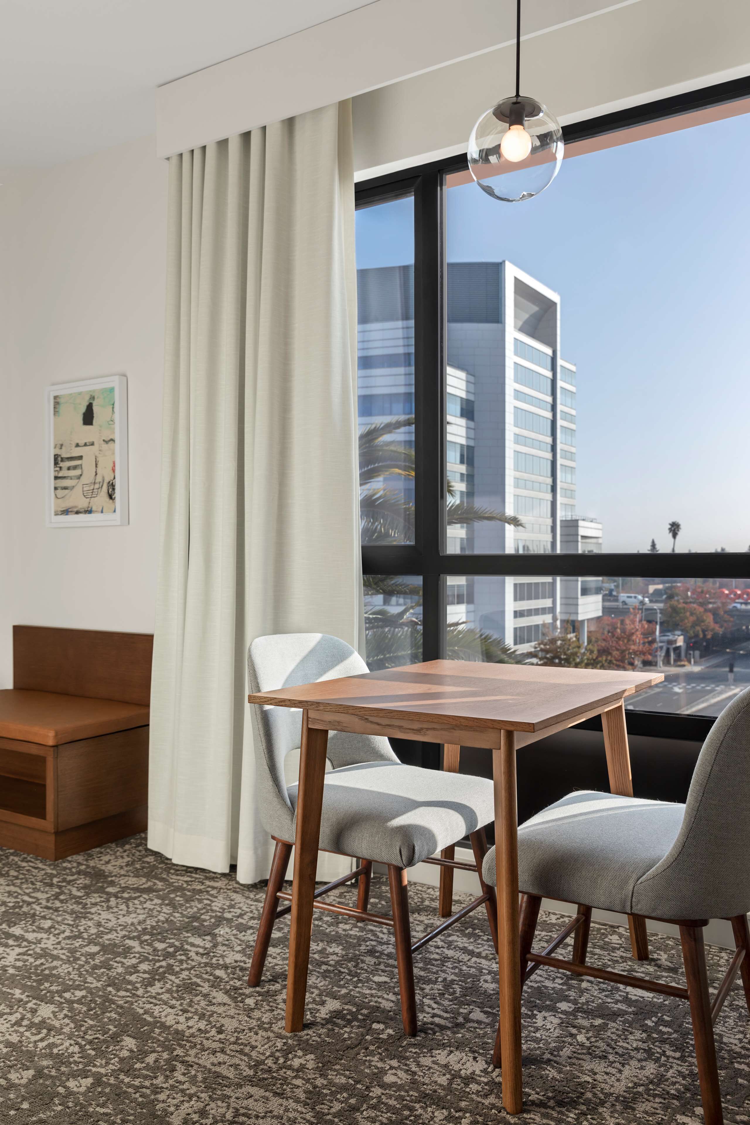 Una habitación de hotel con un ventanal grande, una mesa de madera, dos sillas, un sofá y una alfombra estampada.