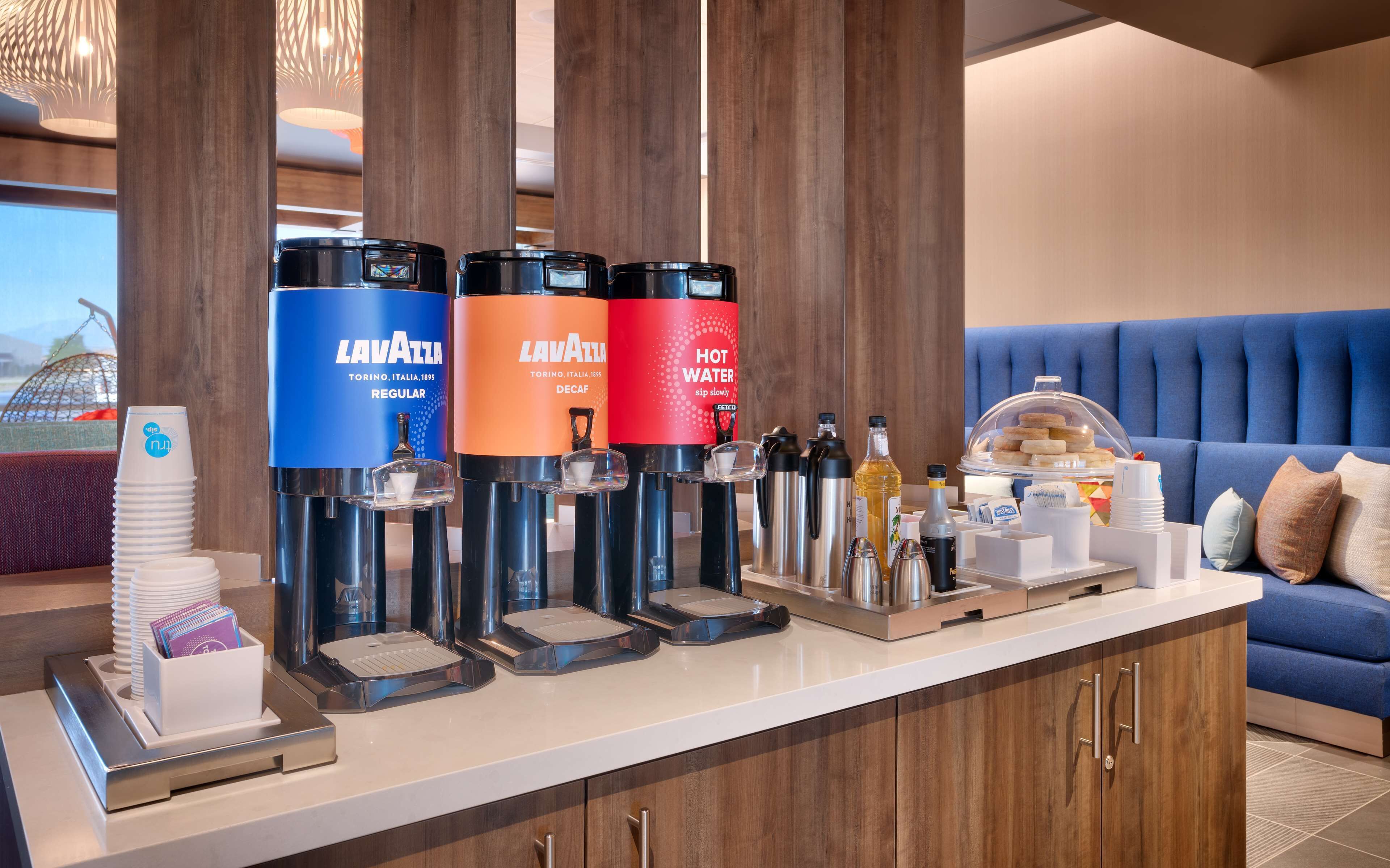 A hotel reception area with a coffee station featuring Lavazza branded dispensers, a buffet with various food items, and a blue upholstered sofa in the background.
