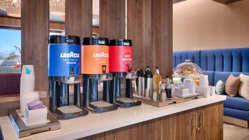 A hotel reception area with a coffee station featuring Lavazza branded dispensers, a buffet with various food items, and a blue upholstered sofa in the background.