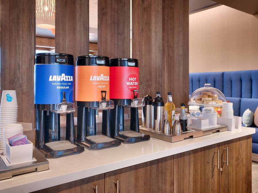 A hotel reception area with a coffee station featuring Lavazza branded dispensers, a buffet with various food items, and a blue upholstered sofa in the background.