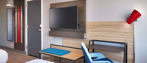 Habitación de hotel con cama, escritorio, silla, televisor montado en la pared y una lámpara roja.