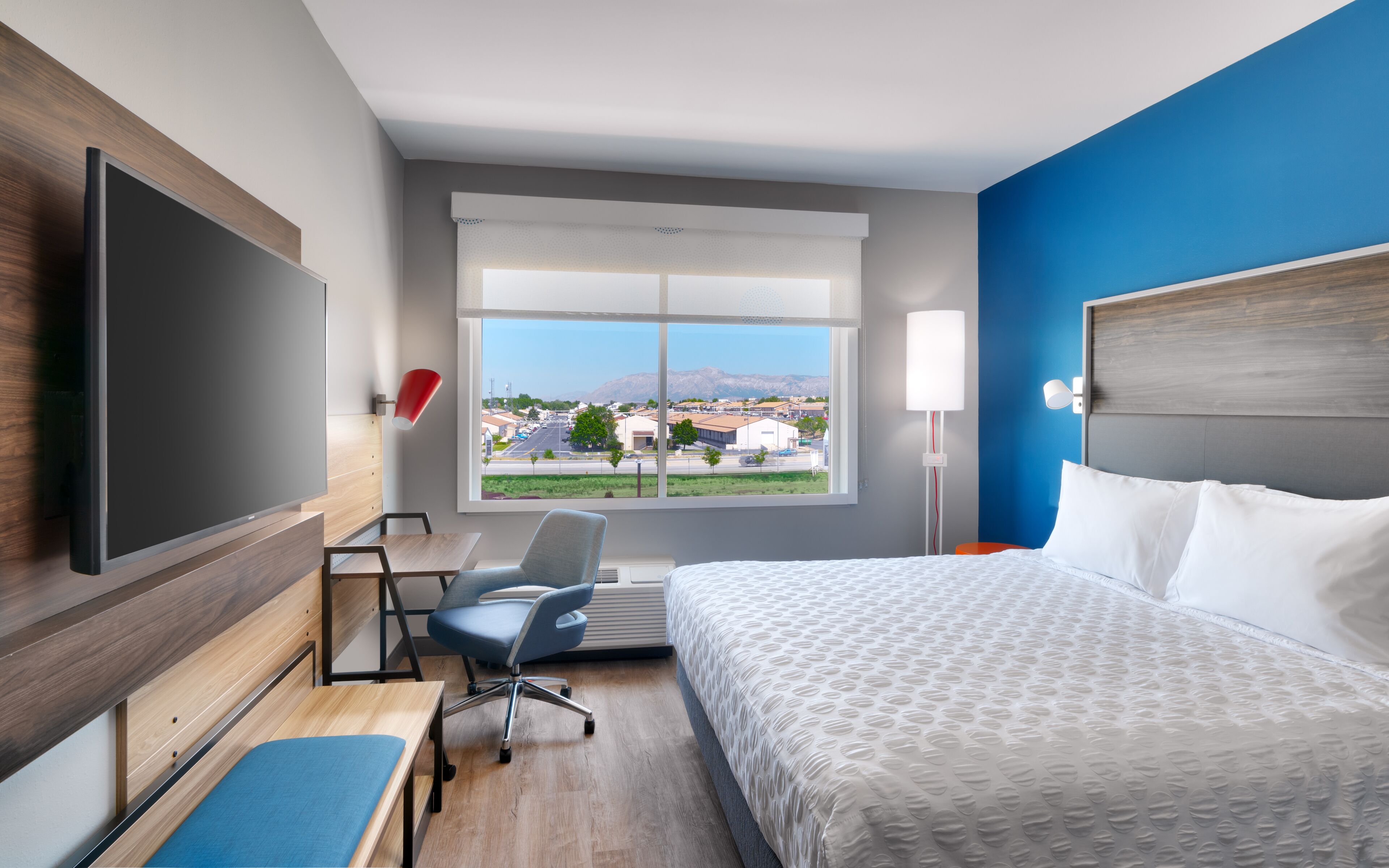 A hotel room with a bed, desk, chair, TV, window, and lamp.