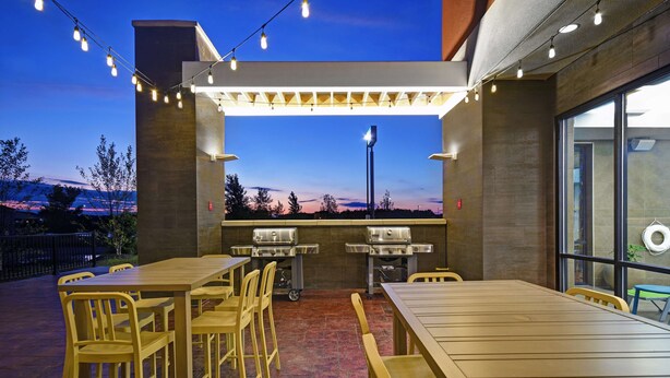 An outdoor patio with wooden tables and chairs, string lights, and a view of the mountains. - Home2 Suites by Hilton Utica, NY (Utica)