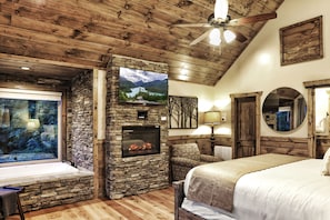 A cozy room with a stone fireplace, a large window, a bed, and a ceiling fan.