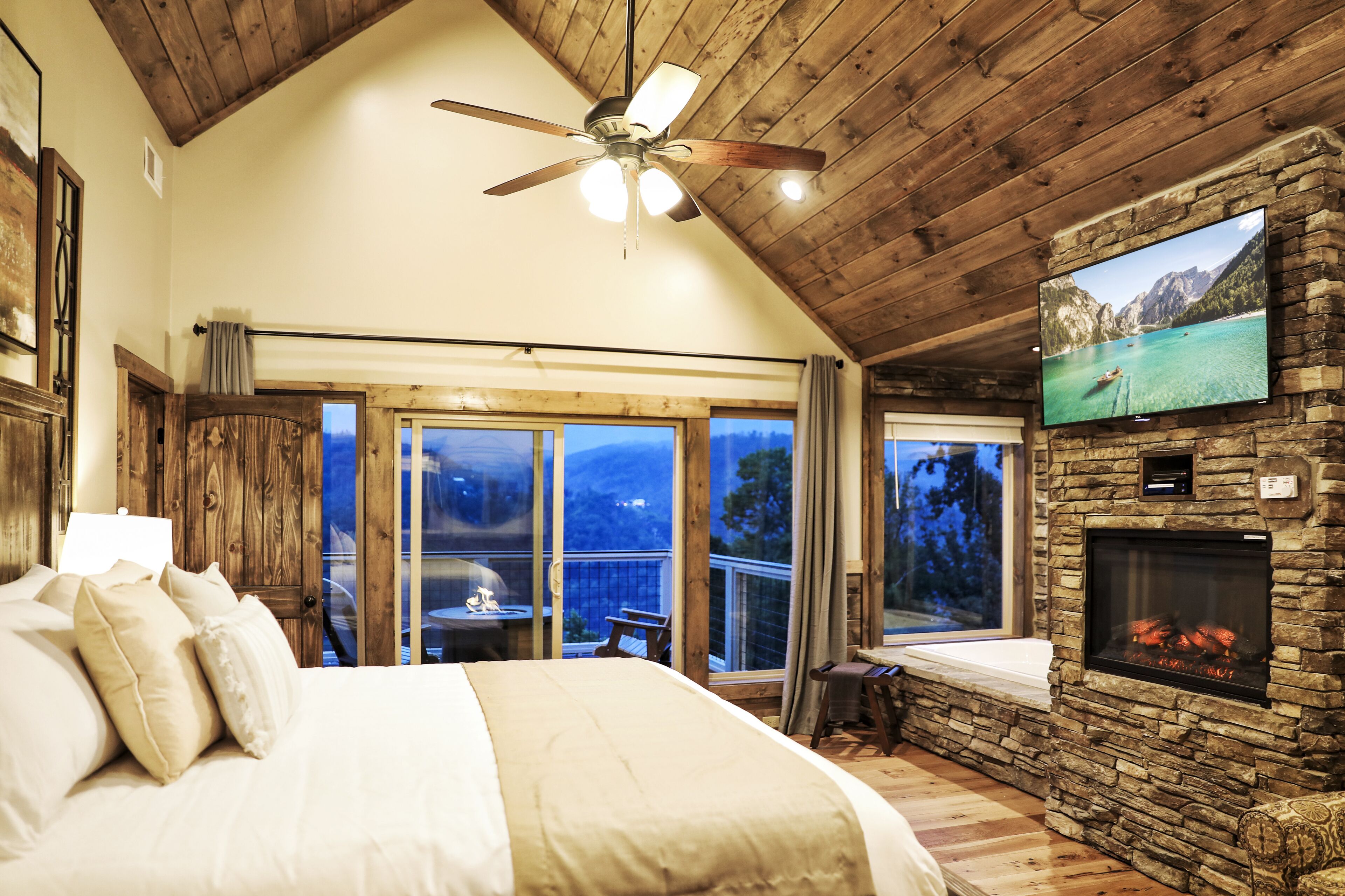 A bedroom with a large bed, a ceiling fan, a stone fireplace, and a TV mounted on the wall.