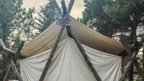 Une tente avec une structure en bois et des parois en tissu blanc, une terrasse en bois, deux chaises bleues et une personne assise sur la terrasse.