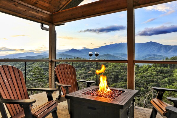 Une terrasse en bois avec deux fauteuils Adirondack, un foyer avec des flammes et une vue panoramique sur les montagnes.