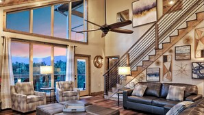 A spacious living room with a large window, a ceiling fan, a staircase, and various artworks.