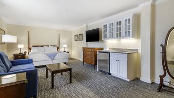 A hotel room with a bed, a sofa, a small table, a desk, a kitchenette, and a TV mounted on the wall.