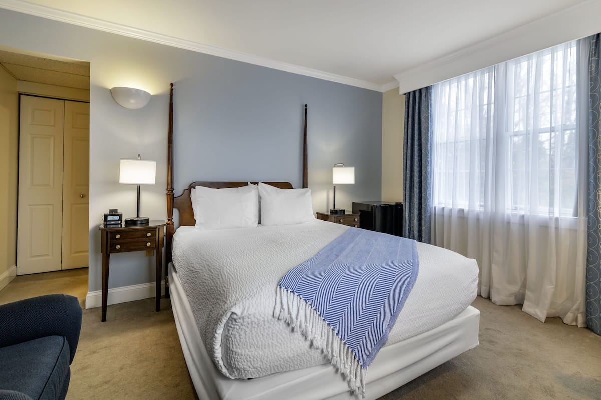 Una cama bien hecha con edredón blanco y una manta de rayas azules y blancas. Hay dos mesitas de noche con lámparas, un tocador de madera y una puerta a la izquierda.
