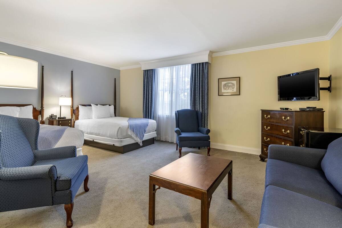 Habitación de hotel con cama, dos sillones, sofá, mesa de centro de madera, cómoda, televisor y ventana con cortinas.
