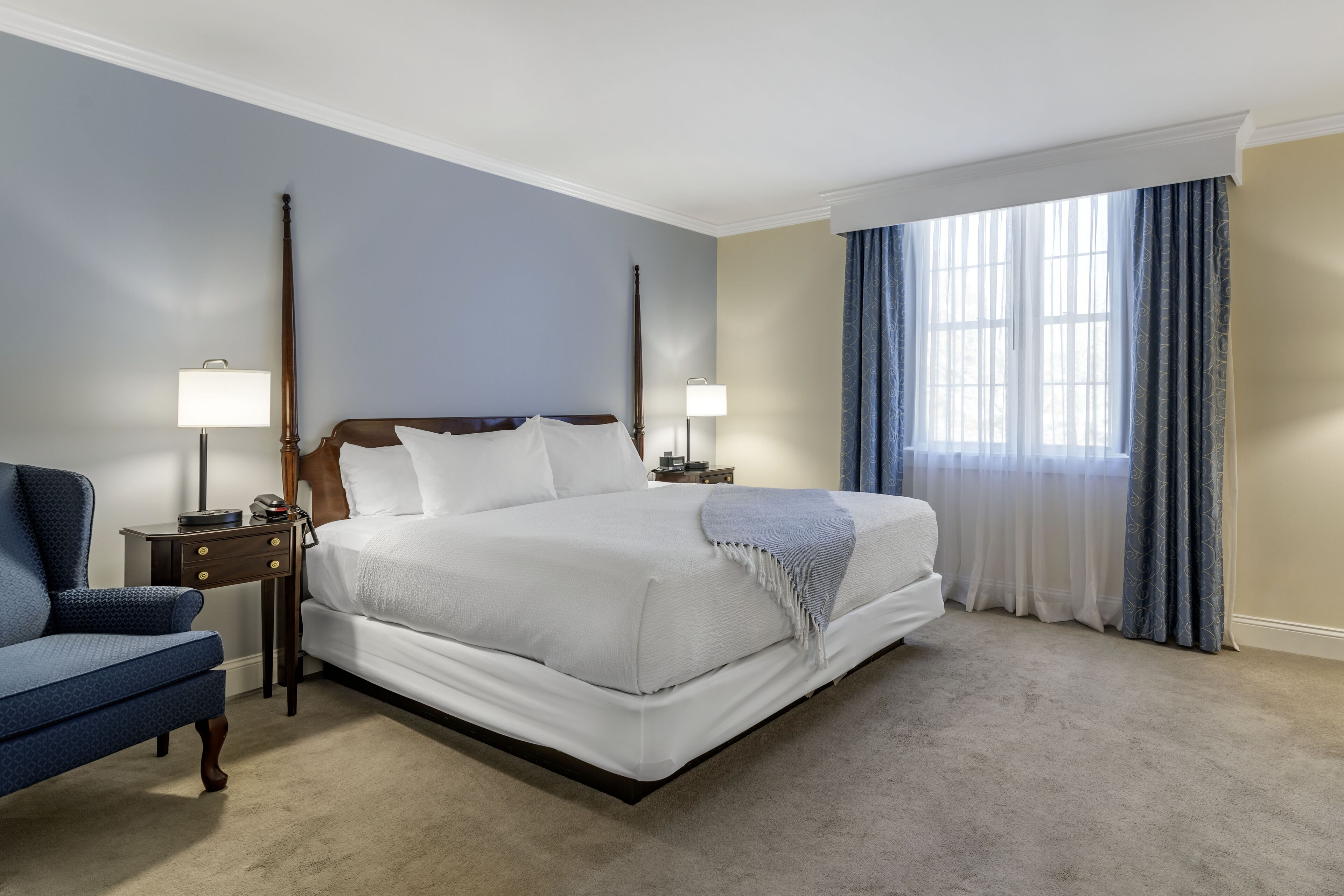 Una habitación de hotel con una cama grande, dos mesitas de noche, una silla y un ventanal con cortinas.