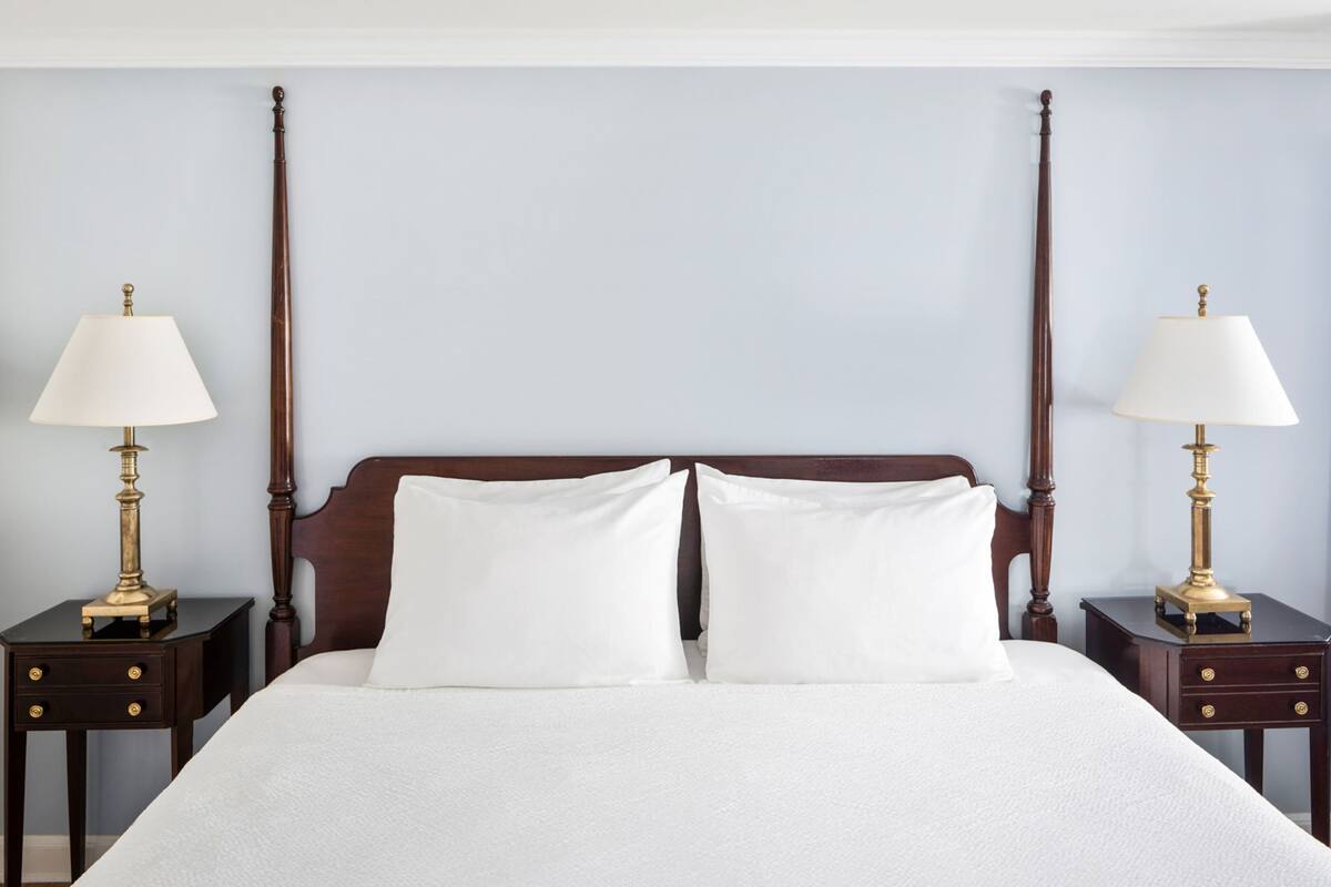 Una cama bien hecha con sábanas blancas, flanqueada por dos mesitas de noche con lámparas, frente a una pared lisa.