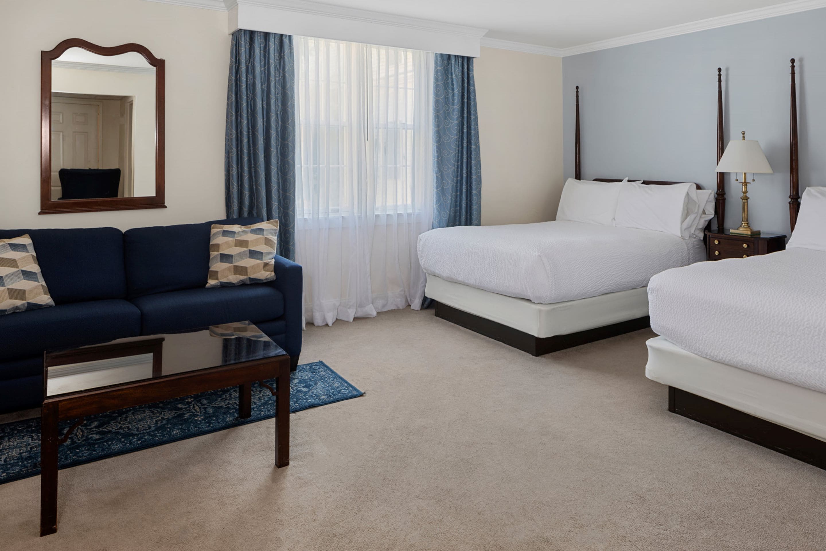 A hotel room with a bed, a sofa, a coffee table, a nightstand, a lamp, a mirror, and a window with curtains.