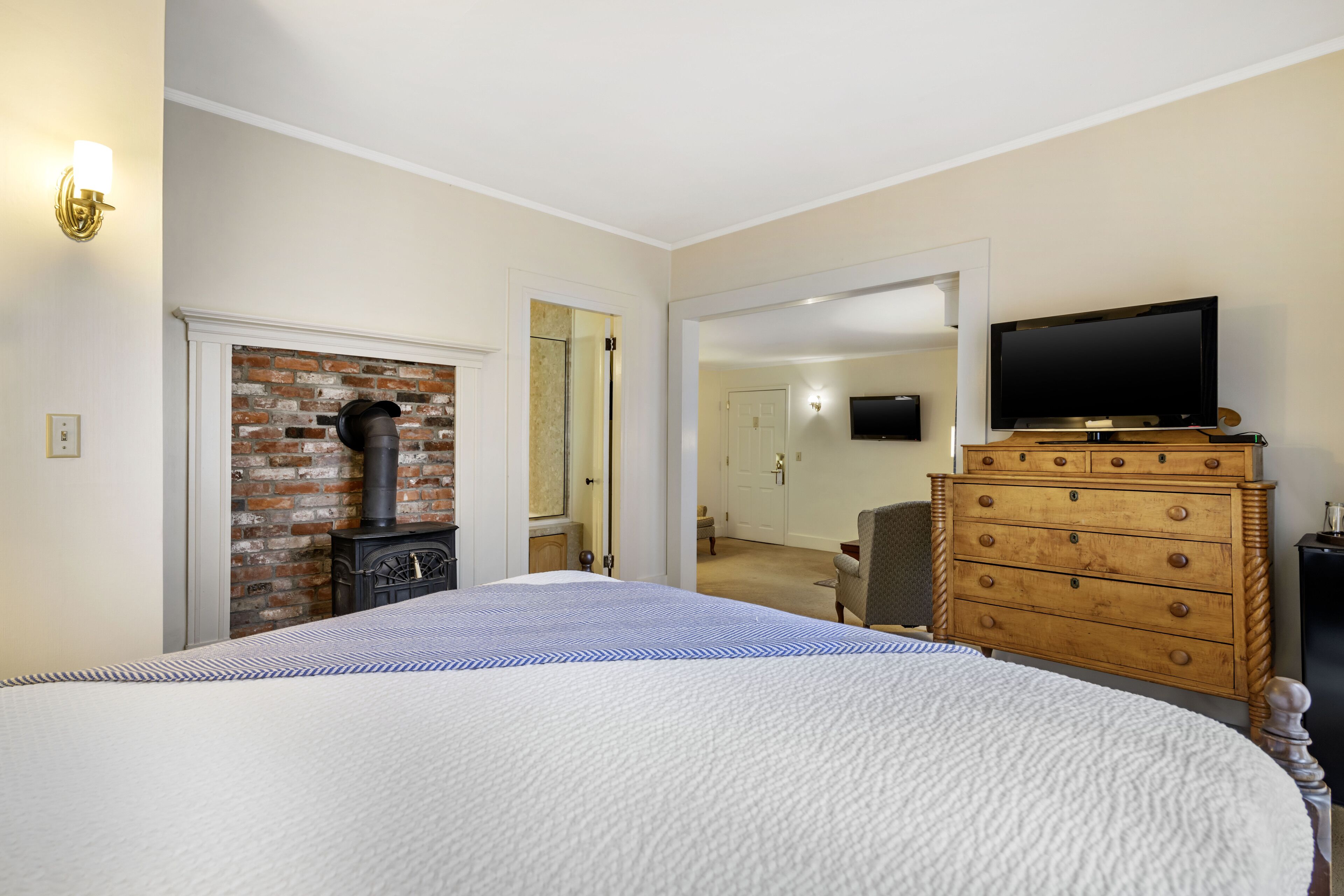 A bedroom with a large bed, a wooden dresser, a television on top, and a brick fireplace.