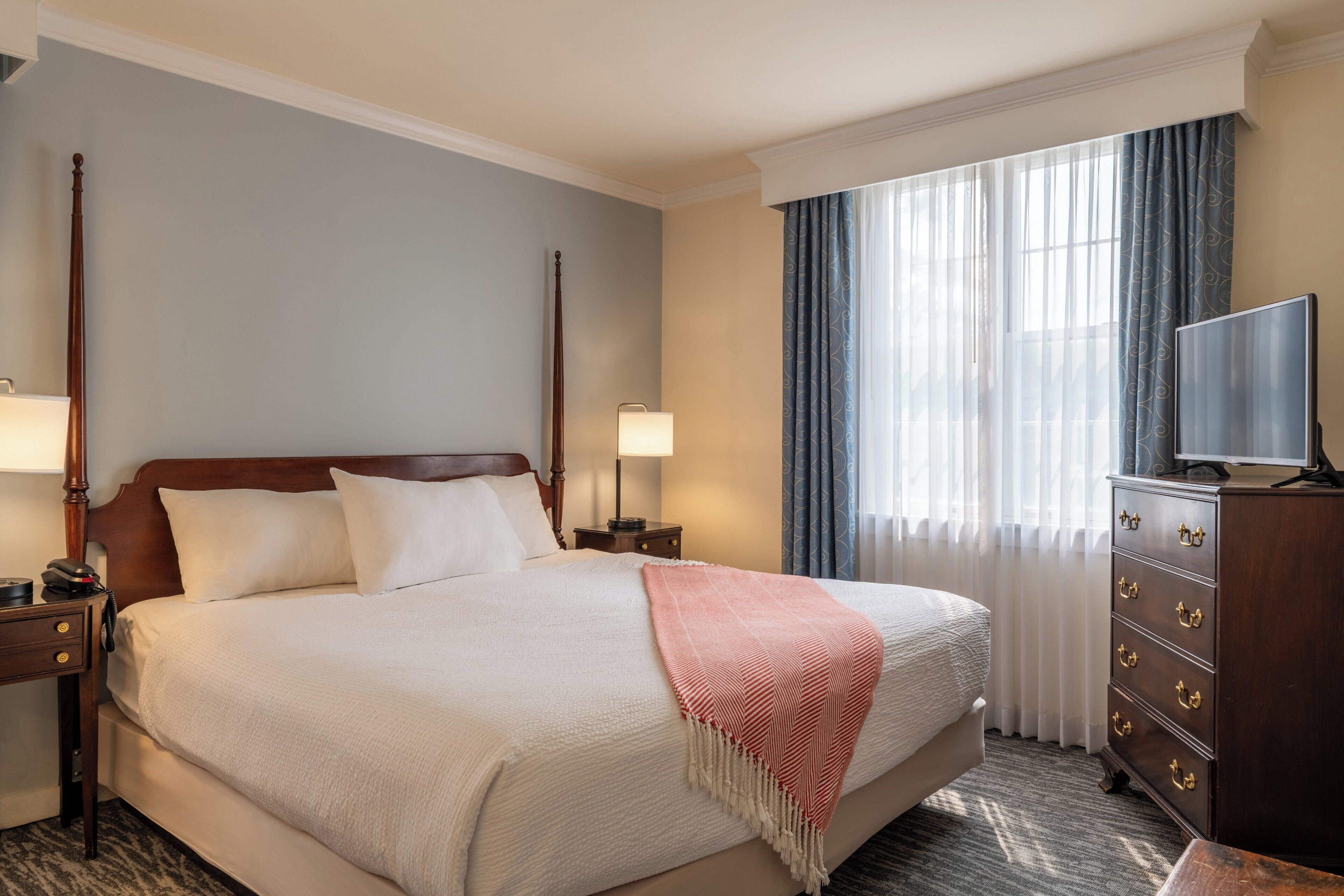 A hotel room with a large bed, two bedside tables, a dresser, a television, and a window with curtains.
