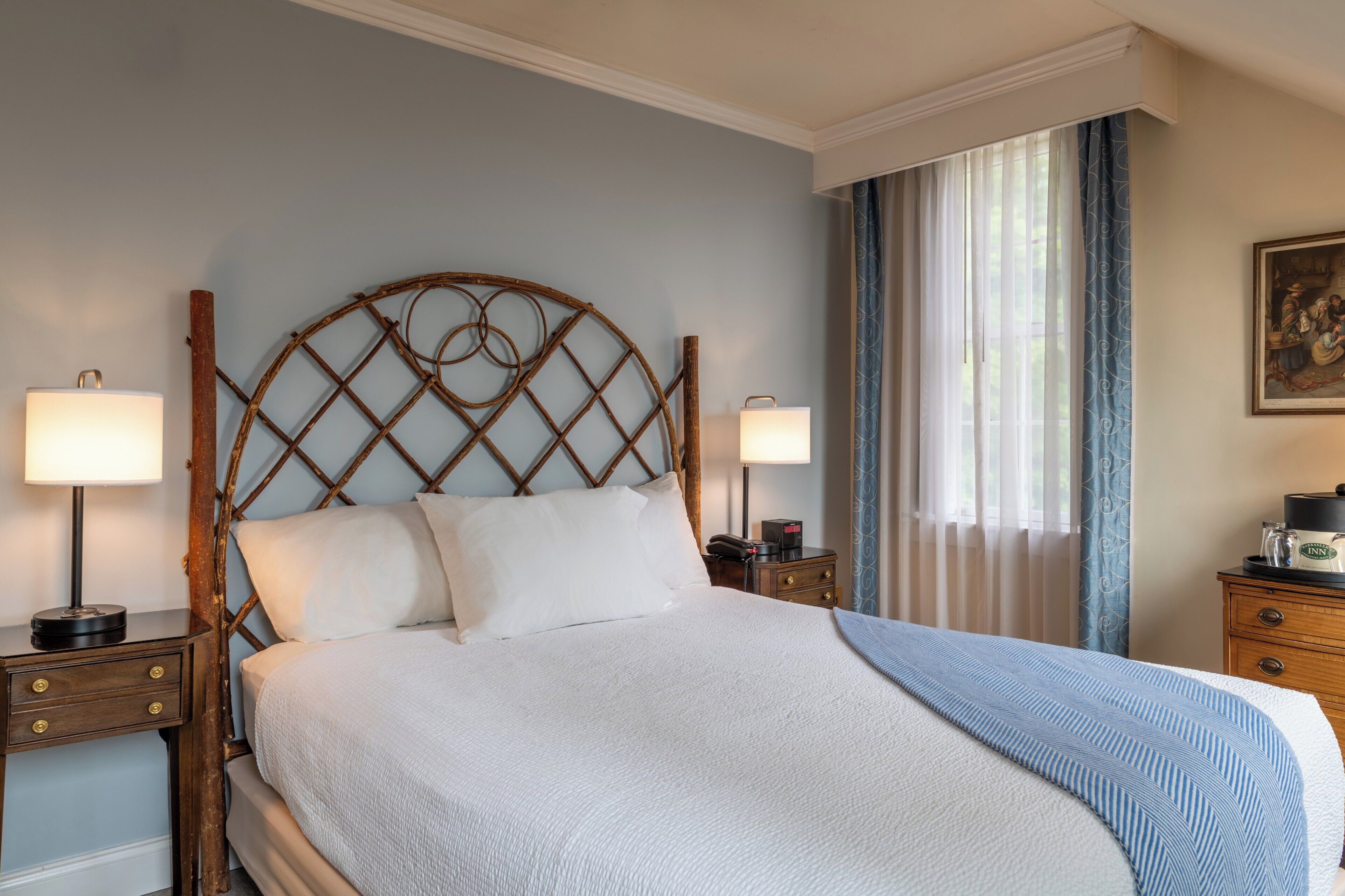 A neatly made bed with a wooden headboard, two bedside tables with lamps, a nightstand with a lamp, and a window with curtains.