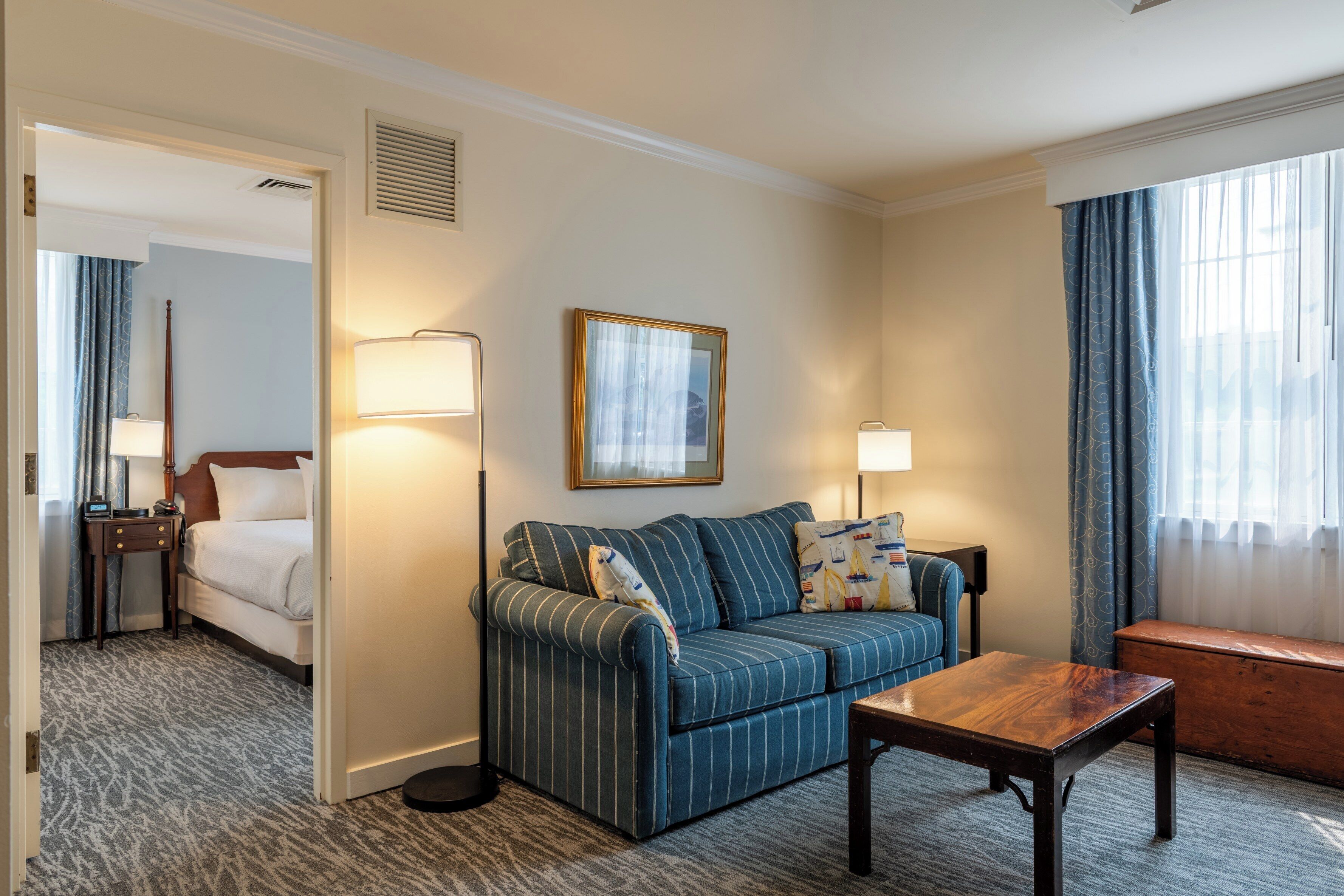 Habitación de hotel con un sofá a rayas, una mesa de centro de madera, una mesita de noche con lámpara, una cama y vistas al exterior a través de una ventana con cortinas.