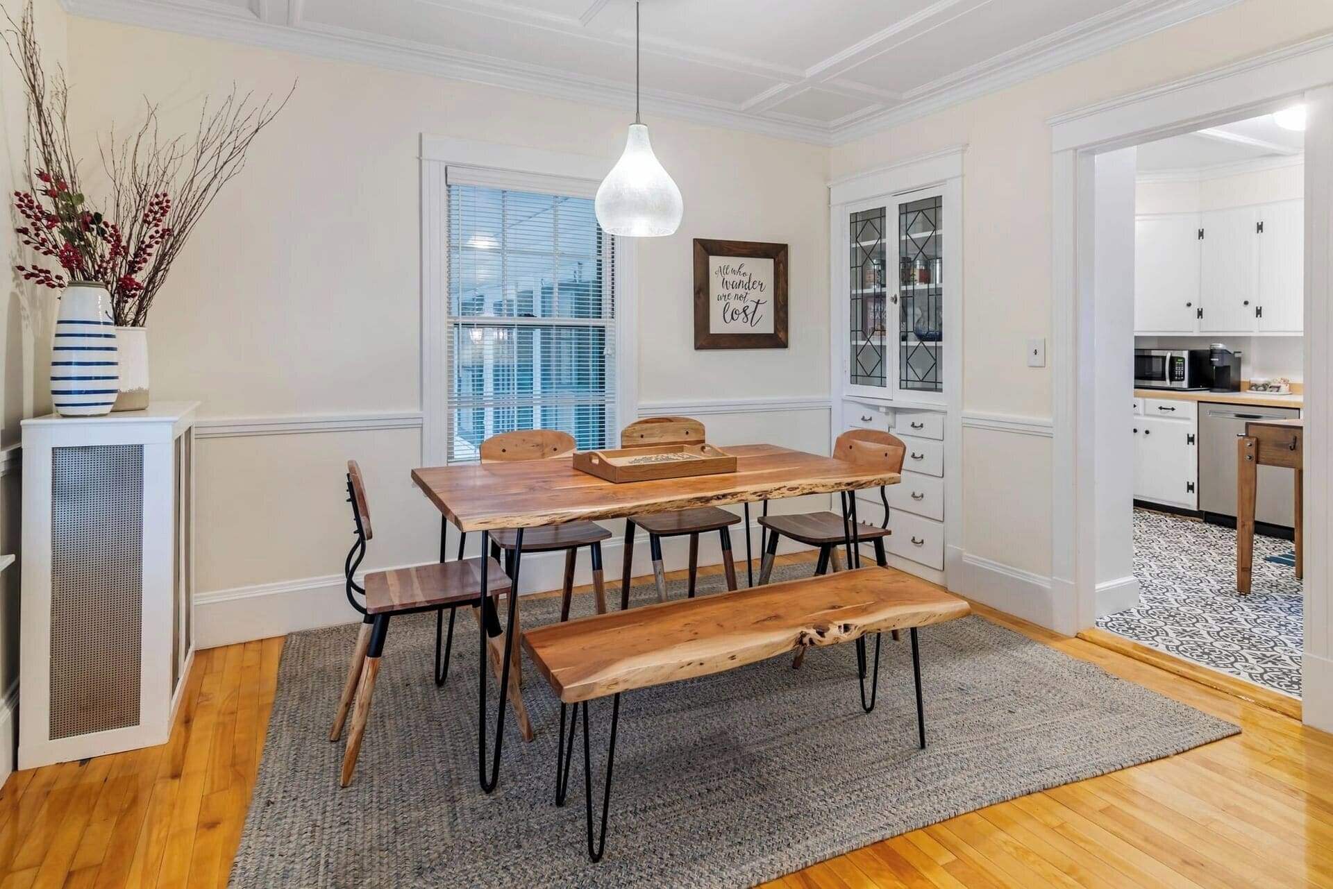 A dining area with a wooden table and benches, a hanging light fixture, a framed inspirational quote, and a view to the kitchen.