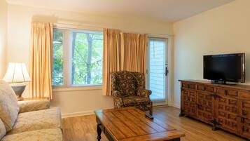 Una sala de estar con un sofá, una mesa de centro, un sillón estampado, un televisor sobre un mueble de madera y una ventana con cortinas.