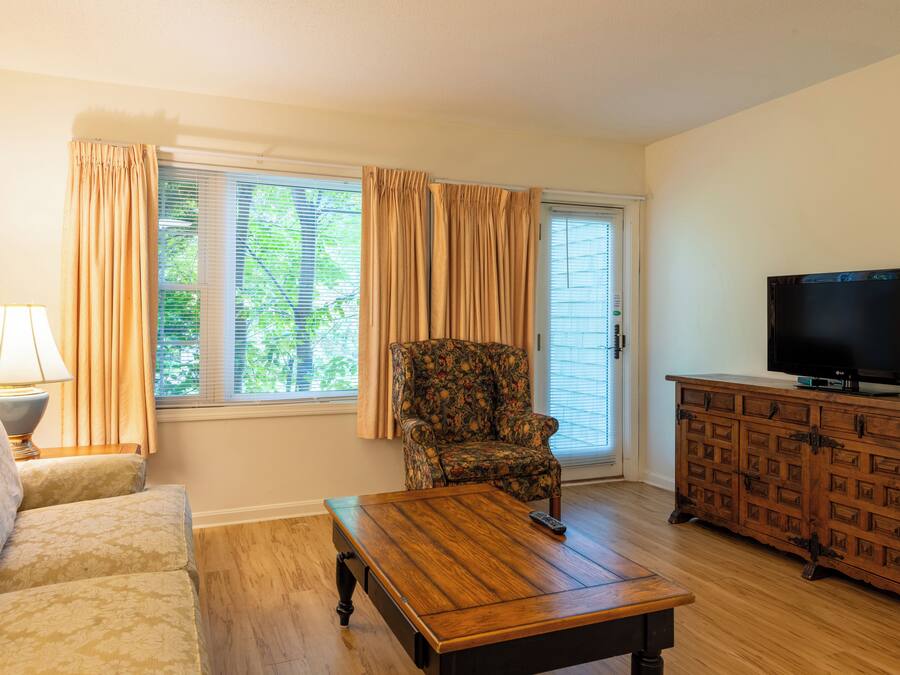 Una sala de estar con un sofá, una mesa de centro, un sillón estampado, un televisor sobre un mueble de madera y una ventana con cortinas.