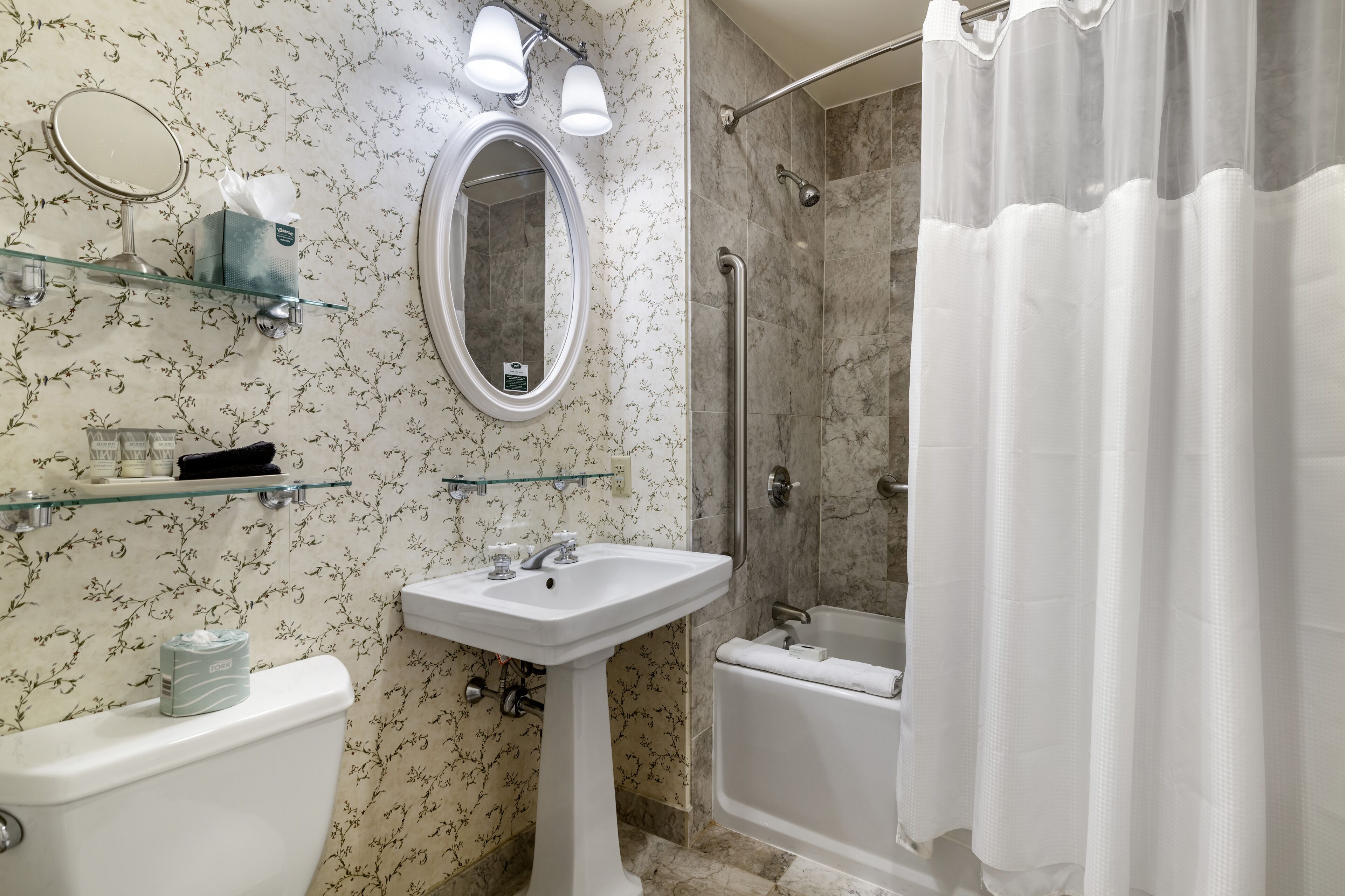 Un baño con inodoro, lavamanos pedestal y bañera. Hay repisas de vidrio sobre el lavamanos y una cortina de ducha.