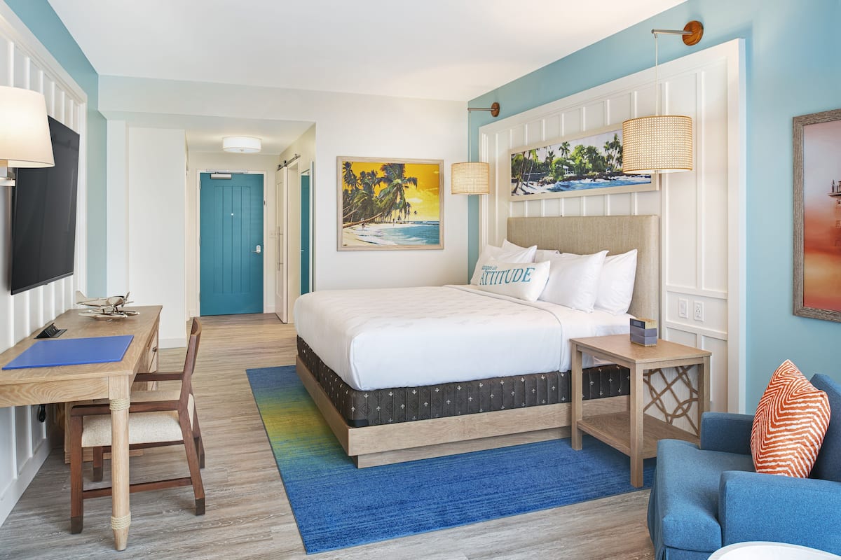 A hotel room with a bed, a desk, a chair, a TV, and a painting on the wall.