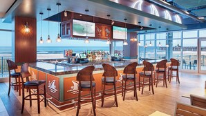 A bar area with a view of the beach, wooden bar stools, and a counter with a menu display.