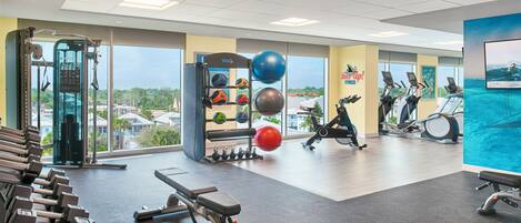 A gym with exercise equipment, a wall with a fitness-related mural, and large windows offering a view of the outdoors.