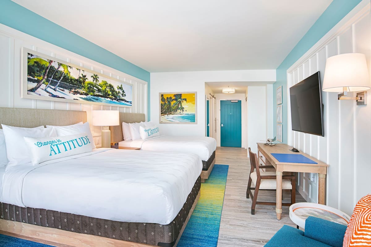 A hotel room with three beds, a desk, a TV, and artwork on the walls.