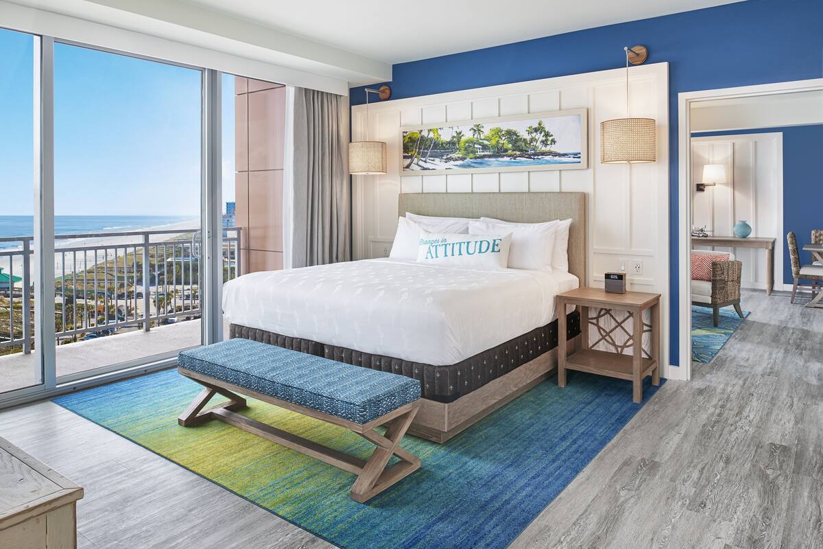A hotel room with a large bed, a bench, a view of the ocean, and a dining area.