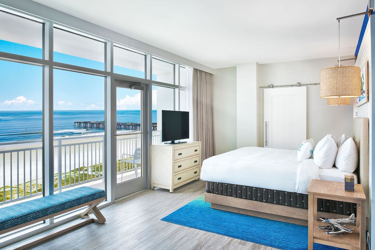 A hotel room with a large window offering a view of the ocean and pier, a bed with a patterned headboard, a bedside table with a lamp, and a bench by the window.