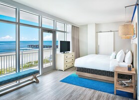 Habitación de hotel con un ventanal amplio que ofrece vistas al océano y al muelle, una cama con cabecera estampada, una mesita de noche con lámpara y un banco junto a la ventana.