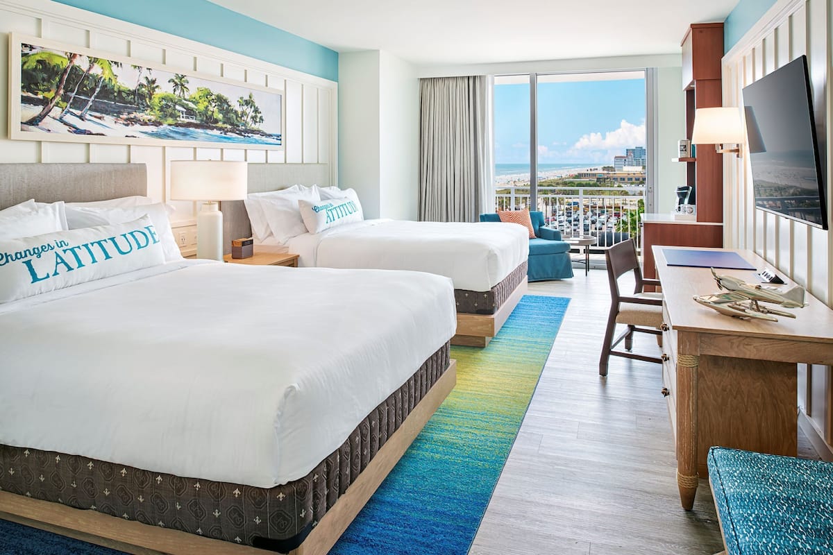 A hotel room with two beds, a desk, a chair, a TV, and a balcony with a view of the beach.