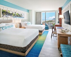 Quarto de hotel com duas camas, uma escrivaninha, uma cadeira, televisão e varanda com vista para a praia.