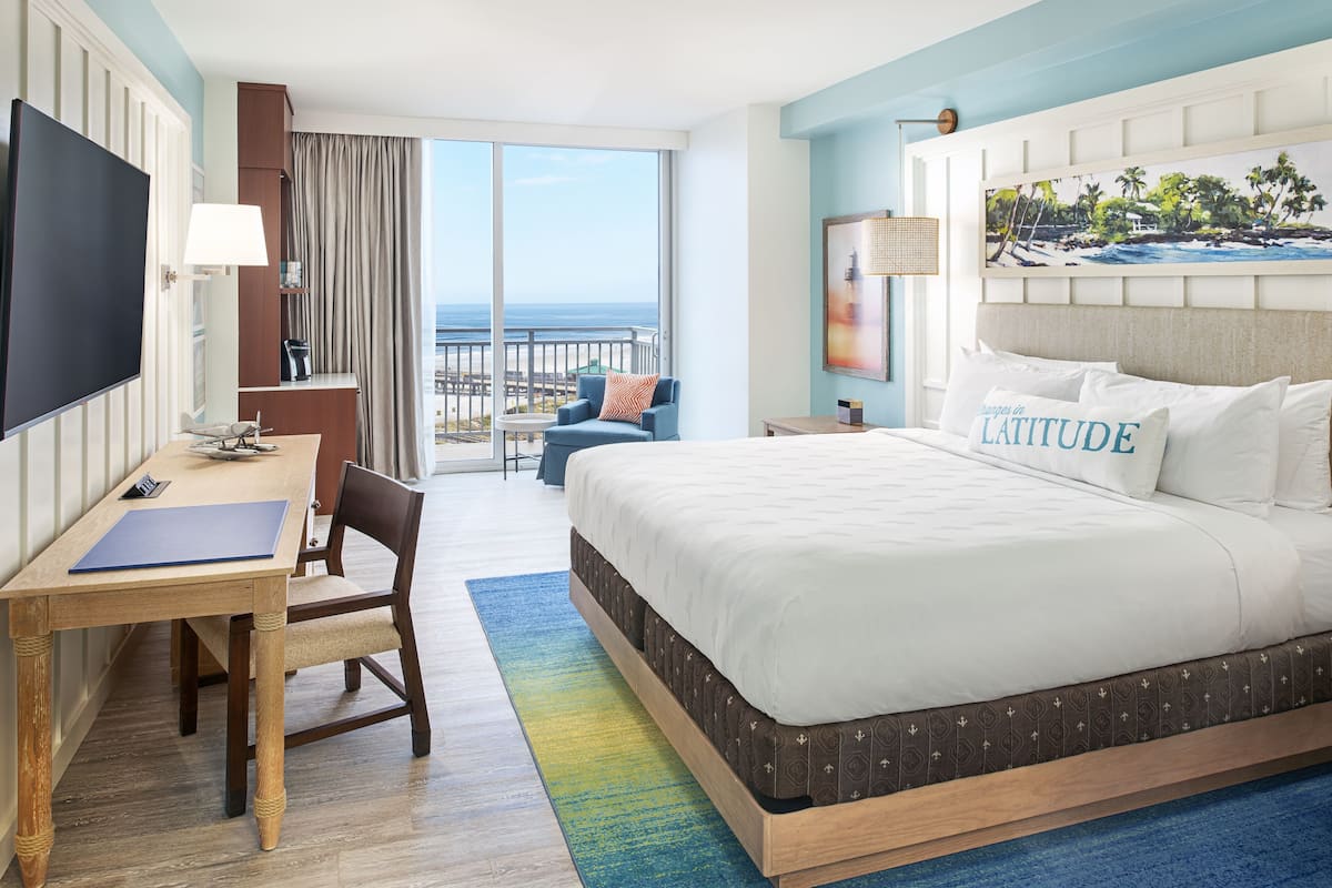 A hotel room with a large bed, a desk with a chair, a TV, and a balcony with a view of the ocean.