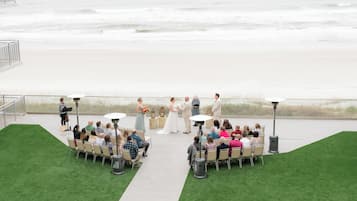 An outdoor wedding ceremony with guests seated on chairs arranged in a semi-circle, facing the couple.