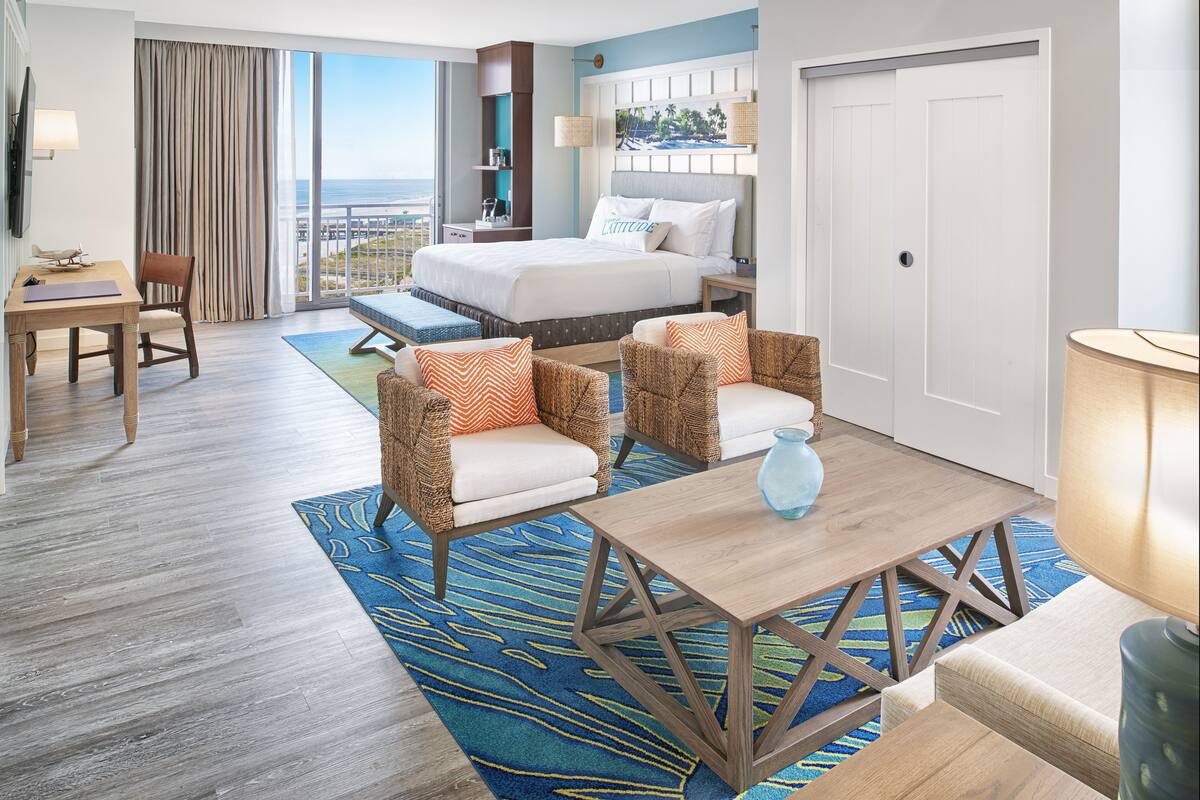 A hotel room with a large bed, a desk, two wicker chairs, a small table, and a view of the ocean.