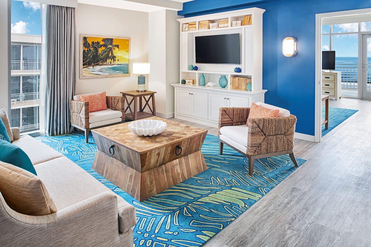 A modern living room with a large TV, comfortable seating, a wooden coffee table, and a view of the ocean.