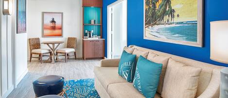 Un soggiorno con una parete blu, un divano con cuscini color acquamarina, un tavolino da caffè e un quadro raffigurante una spiaggia.