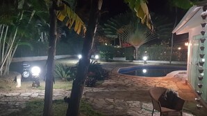 Un sendero de piedra que lleva a una zona de piscina con una silla y una pequeña mesa, rodeado de plantas tropicales y un edificio con luces decorativas.