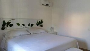 Una cama con ropa de cama blanca, una mesita de noche con lámpara y un aire acondicionado instalado en la pared.