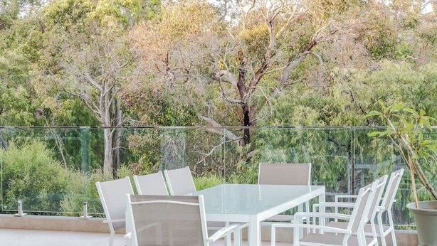 An outdoor patio with a glass table and chairs, surrounded by a glass railing, with trees and greenery in the background. - La Vacanza—Luxe Spacious Home with Ocean Views (Dunsborough)