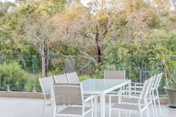 Un patio al aire libre con una mesa y sillas de vidrio, rodeado por una barandilla de vidrio, con árboles y vegetación al fondo.