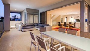 A modern living area with a glass dining table, a sectional sofa, and a kitchen in the background. - La Vacanza—Luxe Spacious Home with Ocean Views (Dunsborough)